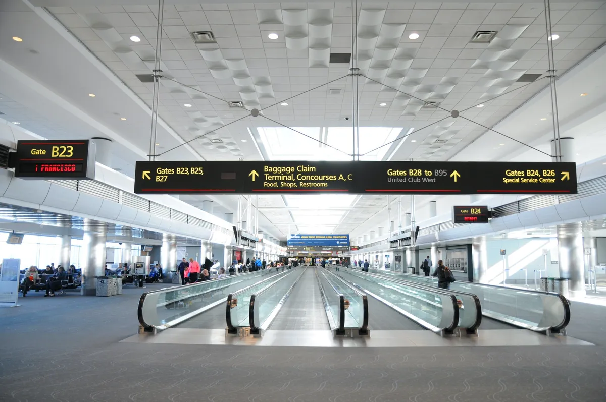 An interior view of a bustling airport terminal with a moving walkway, information boards, and various travelers. The scene evokes the dynamic nature of business travel, highlighting the ease of access to transportation and the seamless movement between work and leisure environments.