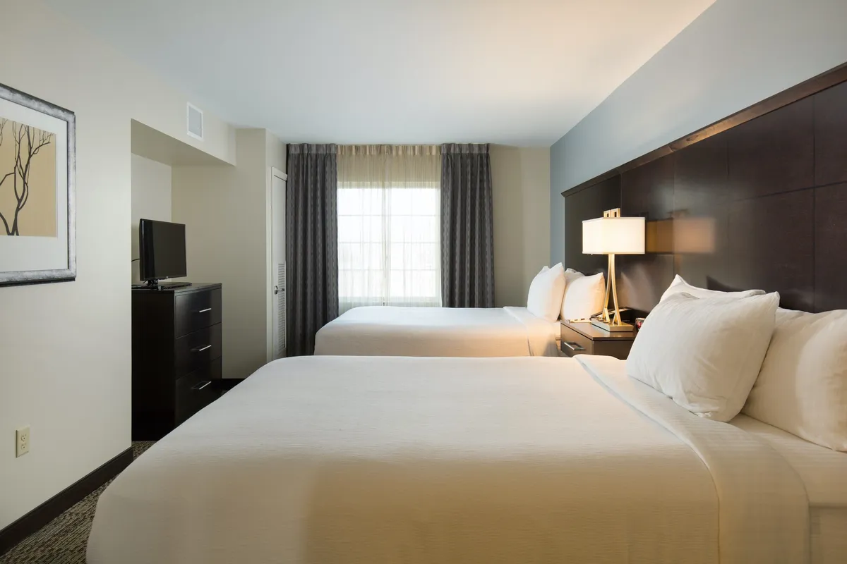The photo displays a spacious bedroom with two queen beds, a large window letting in natural light, a nightstand with a lamp, and a television. The ambiance is relaxed and comfortable, perfect for winding down at the end of the day or enjoying in-room entertainment with family or friends.