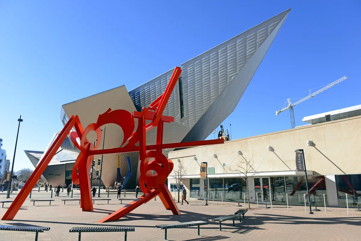 An exterior shot showing a bustling outdoor space with a large red sculpture and a modern architectural building. People are seen walking, socializing, and engaging with public art, creating a dynamic and creative community vibe.