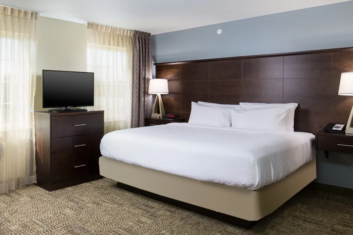 A serene guest room setup featuring a king-size bed, a television, a dresser, and a nightstand with a lamp. The natural light and comfortable furnishings invite relaxation and restful sleep.