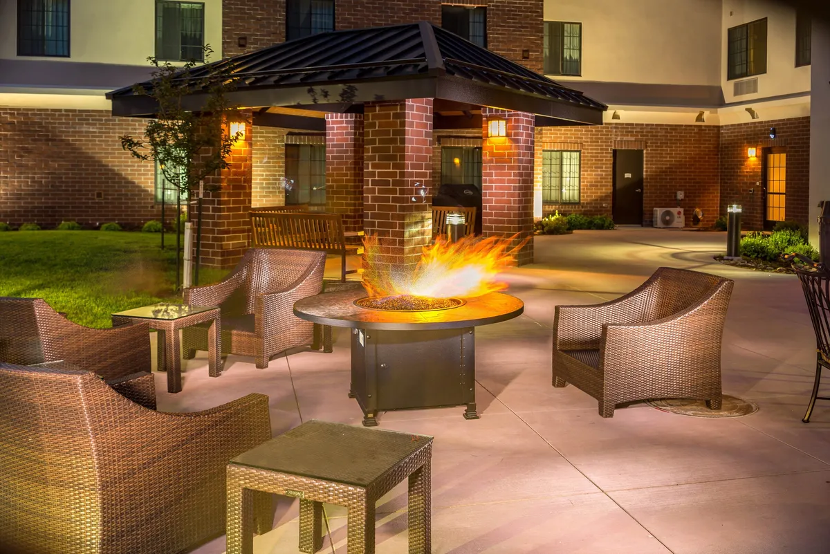 An exterior view of a cozy fire pit area surrounded by seating and attractive landscaping, set against a brick wall. The space is designed for evening conversations, social gatherings, and relaxation under the stars.