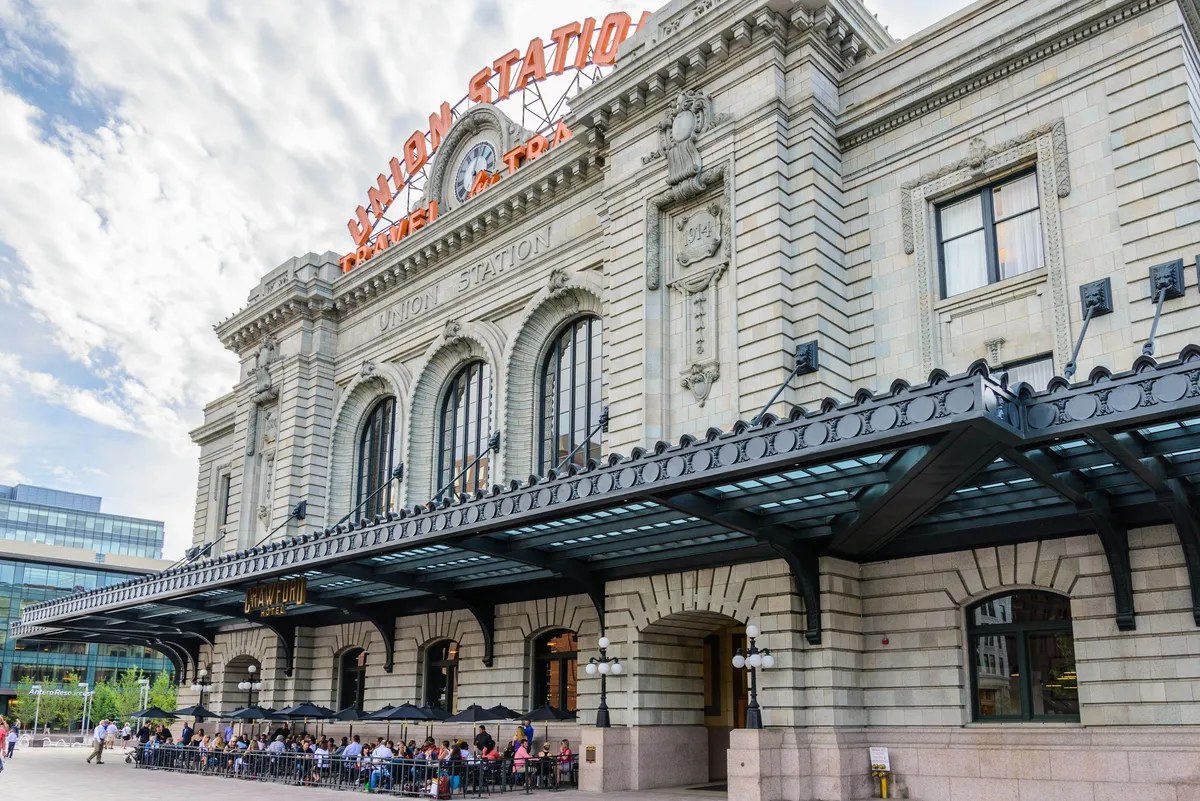 This exterior image captures the grand facade of Denver's Union Station, a historic transit hub with a large, bright sign, outdoor seating, and people enjoying the lively, welcoming plaza. The scene exudes the energy of a city adventure spot, perfect for families looking to explore local attractions and enjoy casual dining together.