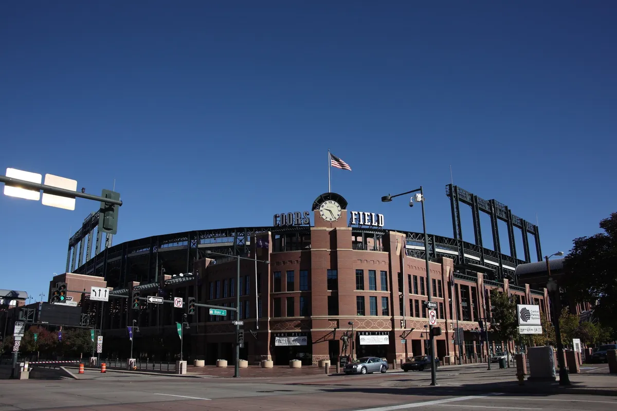 The exterior view of Coors Field, with its recognizable sign, the American flag waving, and distinct architectural details of the stadium. The image captures the energetic atmosphere around this iconic Denver landmark, suggesting opportunities for business travelers to experience local culture and after-hours excitement.