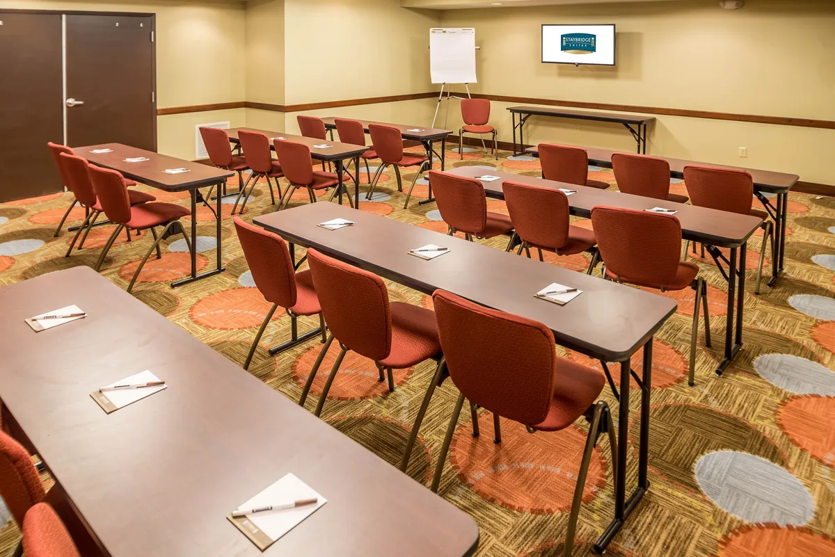 A spacious, well-lit meeting room featuring rows of tables and chairs, a presentation screen at the front, and a whiteboard for brainstorming or presentations. The setup is conducive to meetings, workshops, and networking events.