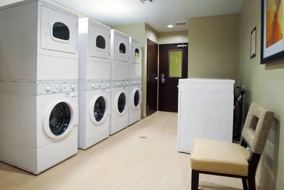 This image depicts a practical and efficient community laundry area, featuring stacked washer and dryer units, a single washing machine, and a nearby seating area where guests can wait or socialize while doing laundry. The scene suggests a shared amenity space designed for convenience and group interaction.