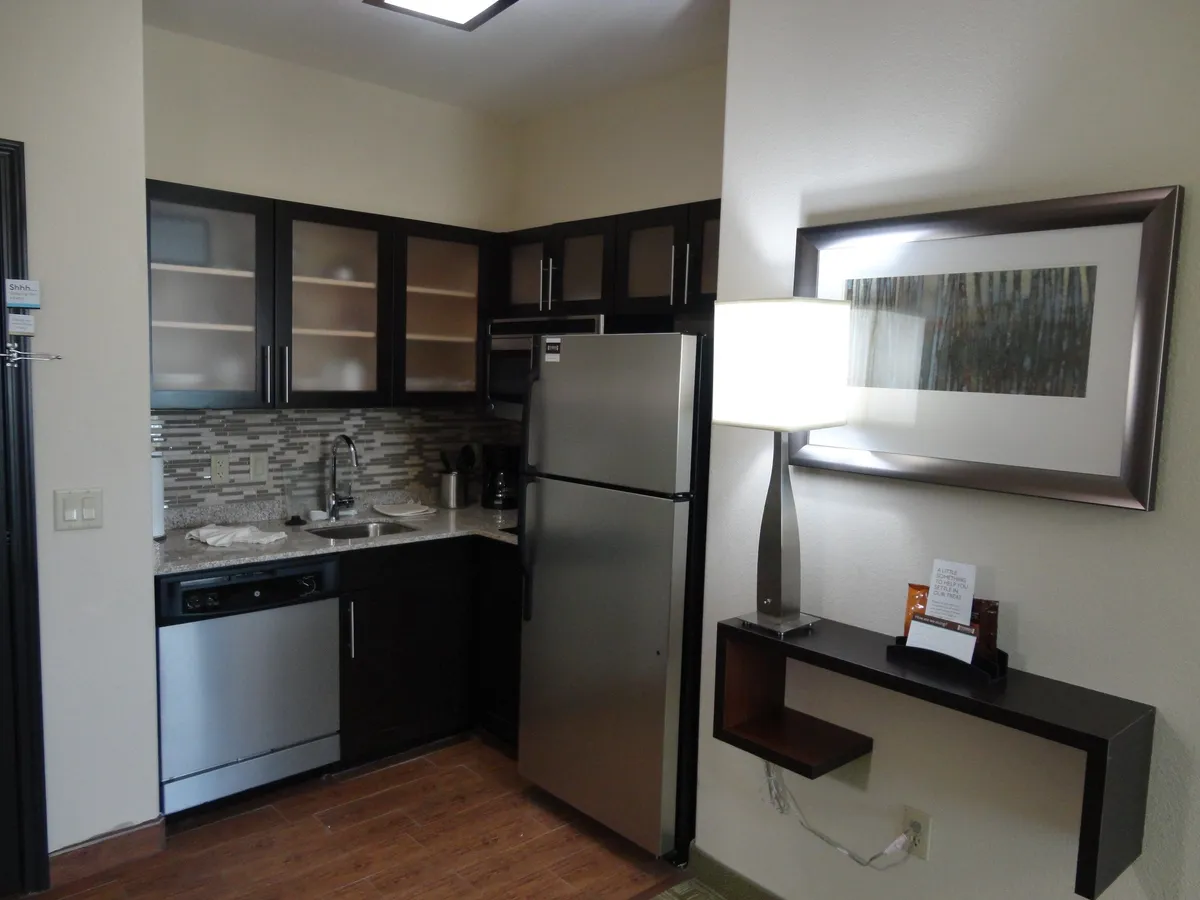 This image showcases a modern kitchen area featuring a stainless steel refrigerator, contemporary cabinetry, and a sleek countertop. The presence of a floor lamp and tasteful wall art adds warmth and a home-like touch, making the space both functional and inviting. The scene hints at an area ideal for cooking, entertaining, or sharing meals.